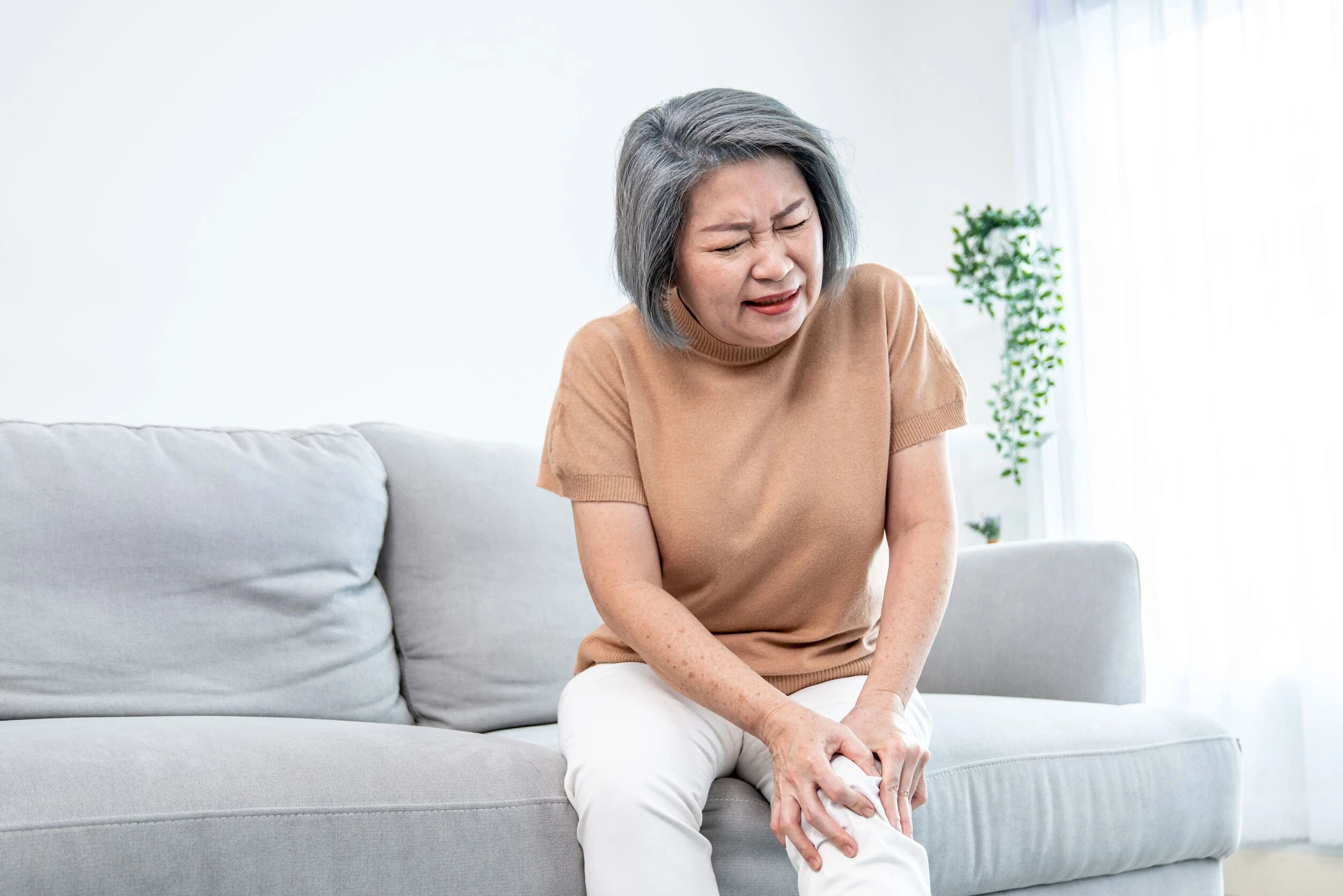 Elderly Woman In Pain Sitting On Sofa Grabbing Her Painful Knee