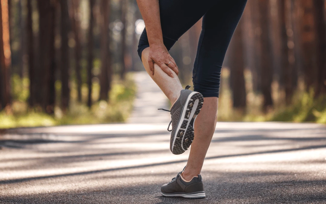 Half Body Woman In Running Shoes Holding On To Her Calf Due To Muscle Cramps In A Park Background